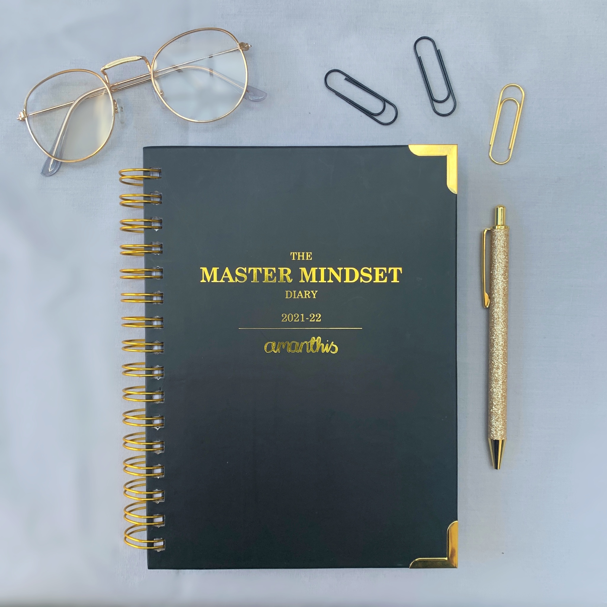 Master Mindset Classic Cover Academic Diary Grey background with black and gold luxury academic planner in the centre. Gold glasses in the top left, paperclips and pen also visible