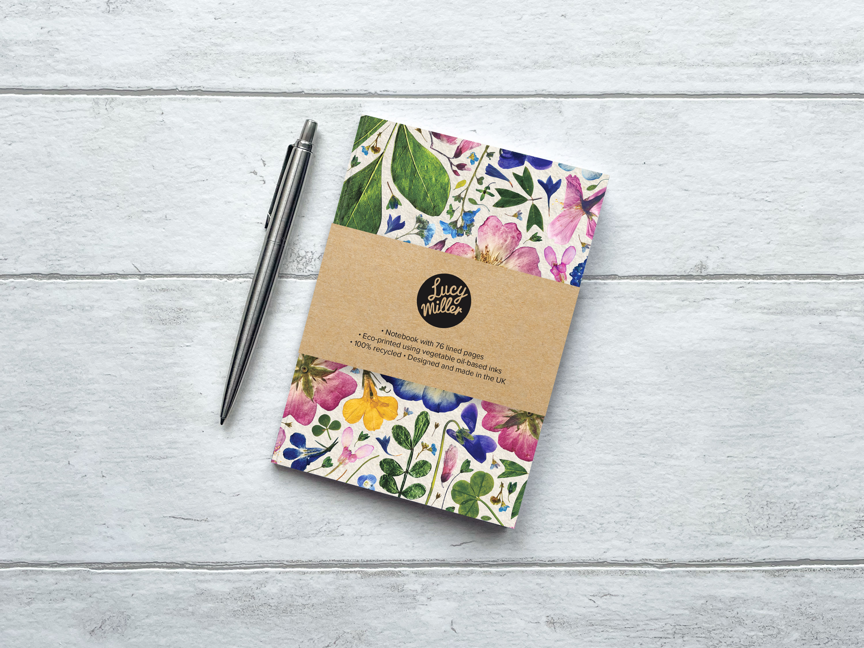 A6 Floral Notebook An A6 recycled notebook with a pressed flower design, on a grey wood background, and a pen to the side. The inside pages are lined..