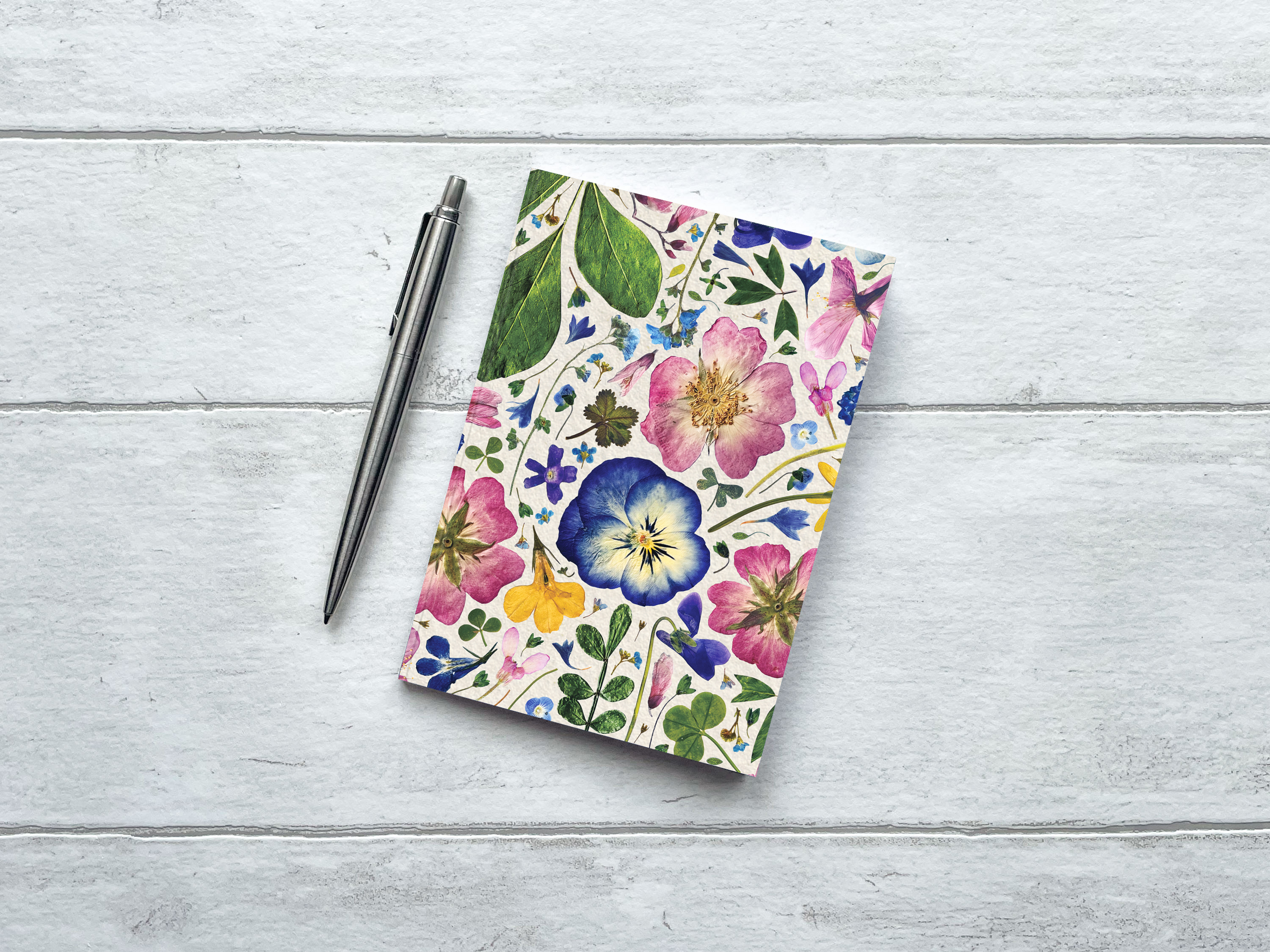 A6 Floral Notebook, Ruled An A6 recycled notebook with a pressed flower design, on a grey wood background, and a pen to the side. The inside pages are lined..