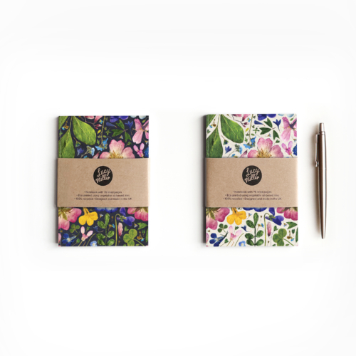 Two A6 recycled notebooks with a pressed flower design. One with a light, natural background. One with a dark, ink background. The notebooks lie on a white background, with a pen to the side.